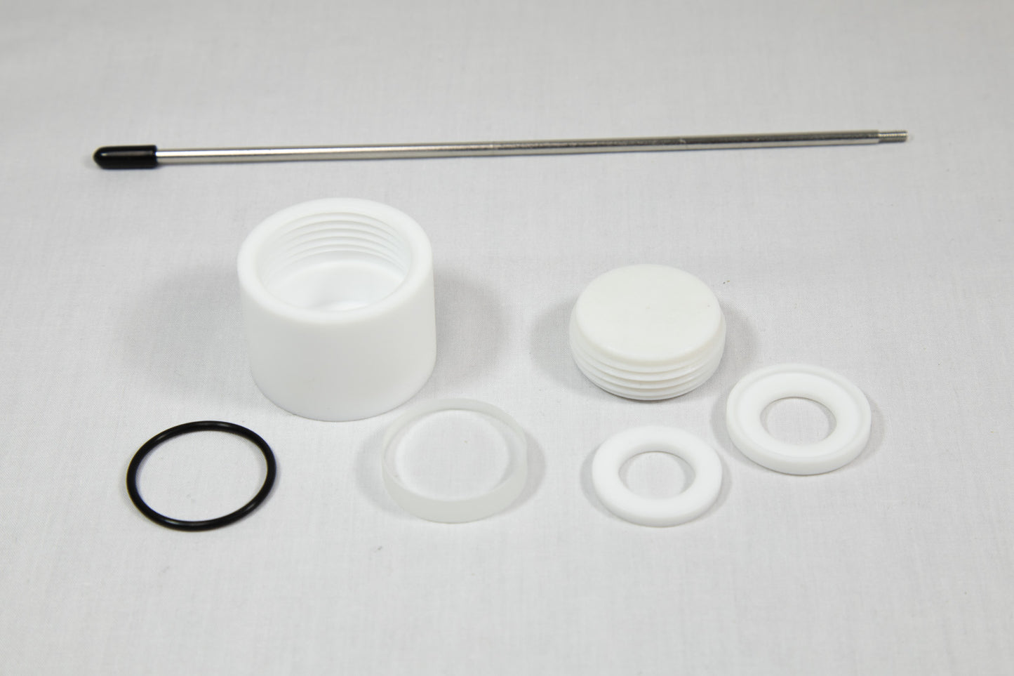 A white plastic valve, a rubber washer, and a metal rod arranged together on a neutral background.