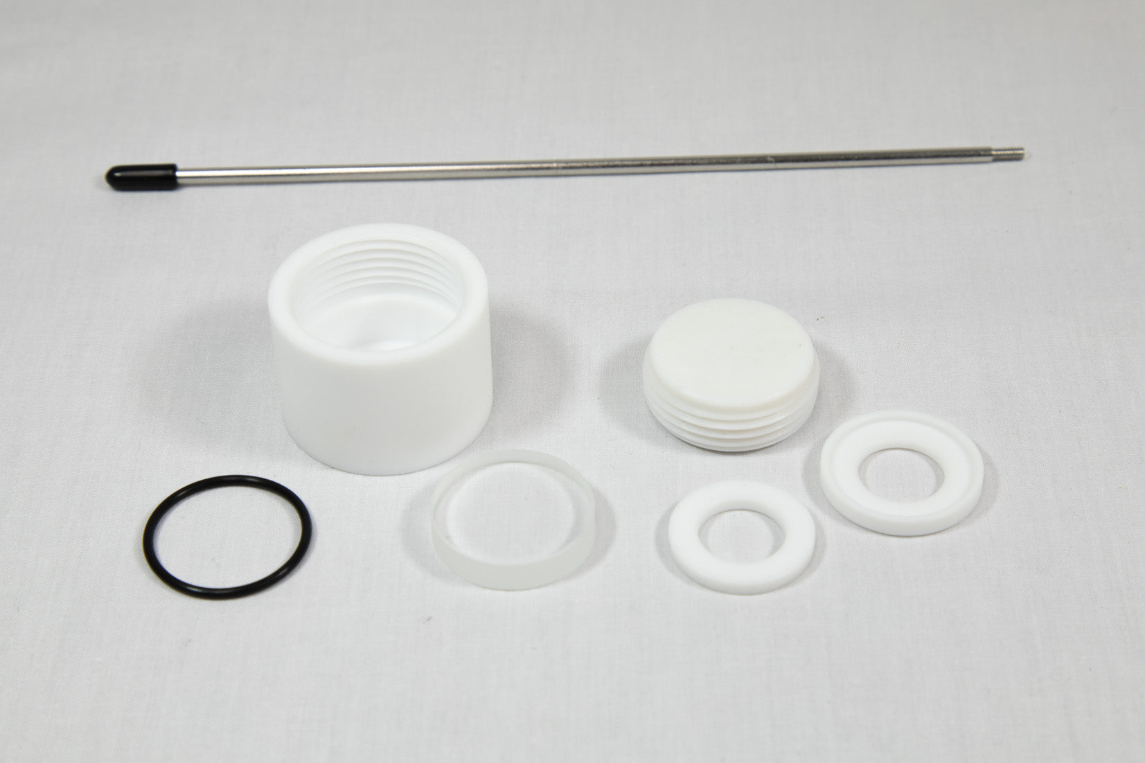 A white plastic valve, a rubber washer, and a metal rod arranged together on a neutral background.